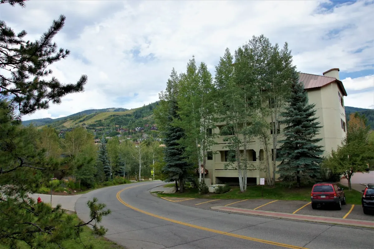 This exterior shot features the building nestled among mature trees with a mountain view in the background. The setting is tranquil, with visible pathways that suggest easy access for walks or biking, and the building's architecture hints at modern comfort within a natural landscape.