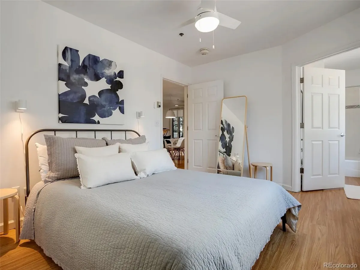 This image features a cozy bed adorned with decorative pillows, abstract artwork on the wall, a floor mirror, and a doorway leading to an adjacent room. Soft morning light fills the space, establishing a serene and inviting mood.