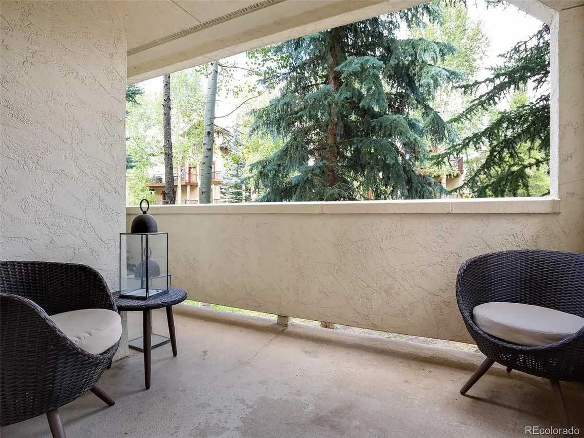 A pair of wicker chairs and a lantern occupy a peaceful outdoor nook with a view of lush trees, suggesting a serene spot for reading, relaxing, or birdwatching. The setting is calm and intimate, ideal for personal moments or shared conversations.