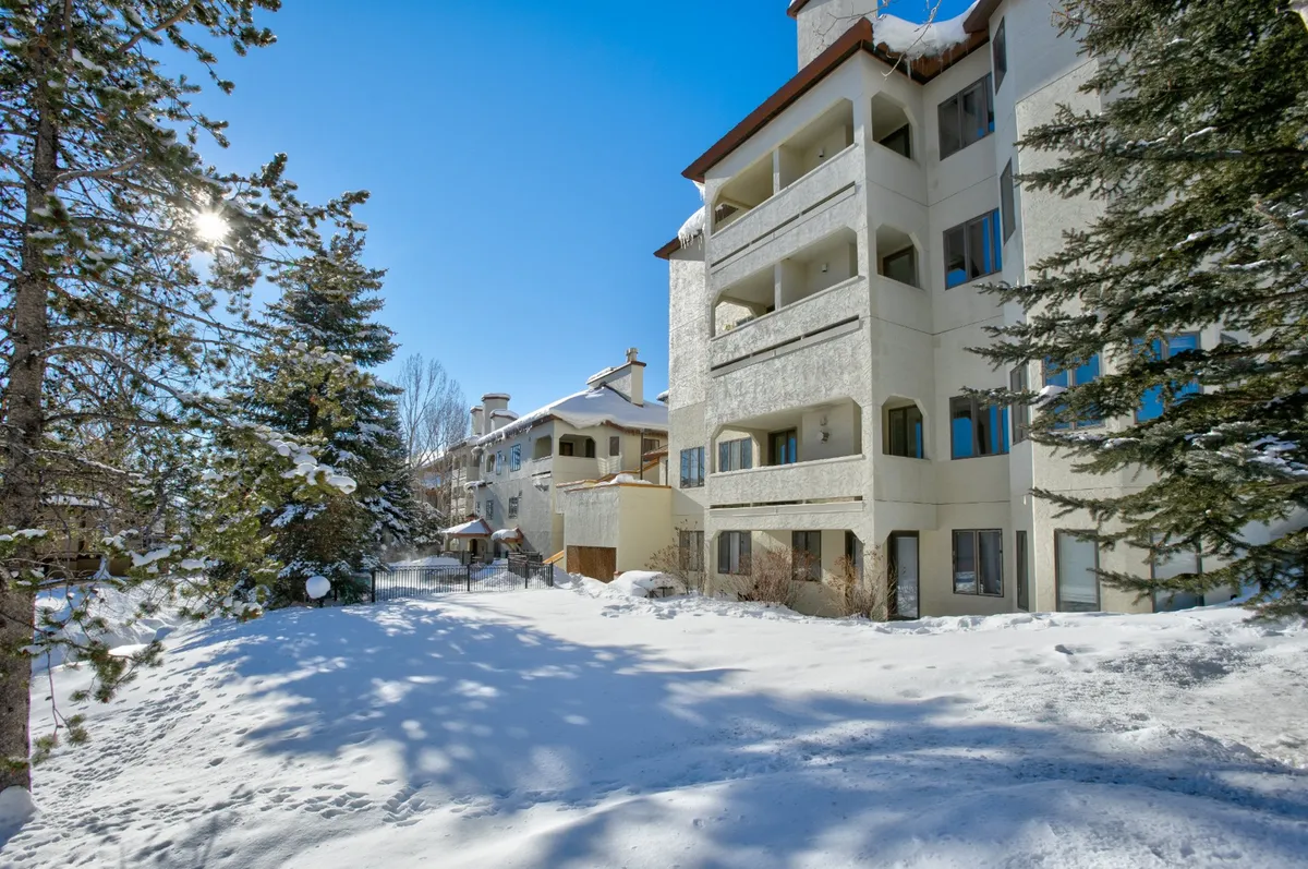 This image features a snow-covered ground surrounding the building facade and trees, capturing the property in a winter setting. The tranquil snow scene directly evokes the ski season atmosphere and the proximity to mountain activities.