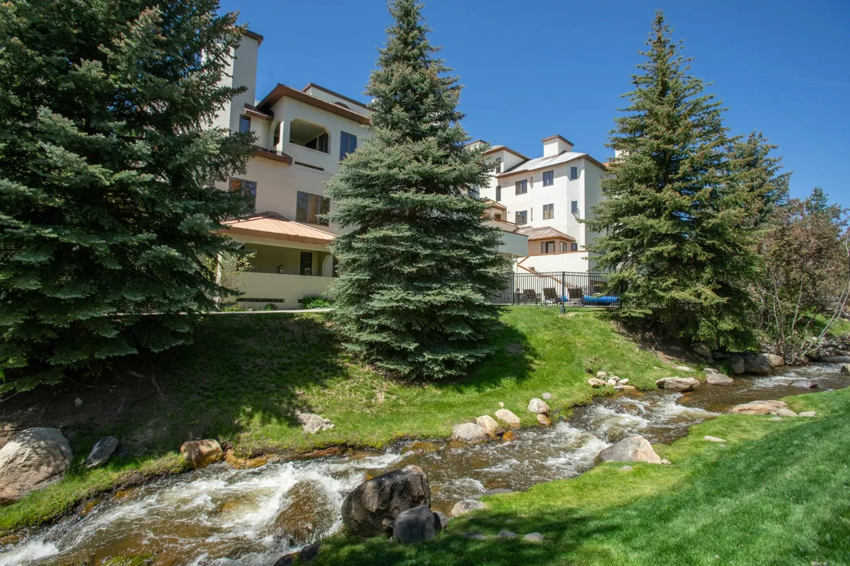 This photo showcases a serene flowing stream bordered by evergreen trees and a view of the building’s facade. The scenery is tranquil, ideal for picnics by the water, taking nature walks, or simply enjoying the peaceful shade. The proximity of the building to these natural features emphasizes accessibility for residents and guests.