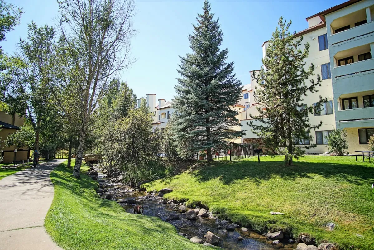 A tranquil creek flows beside manicured lawns and tall evergreen trees, with paths perfect for a stroll. The building exterior is visible, nestled harmoniously in the landscape, inviting exploration of the surrounding natural beauty.