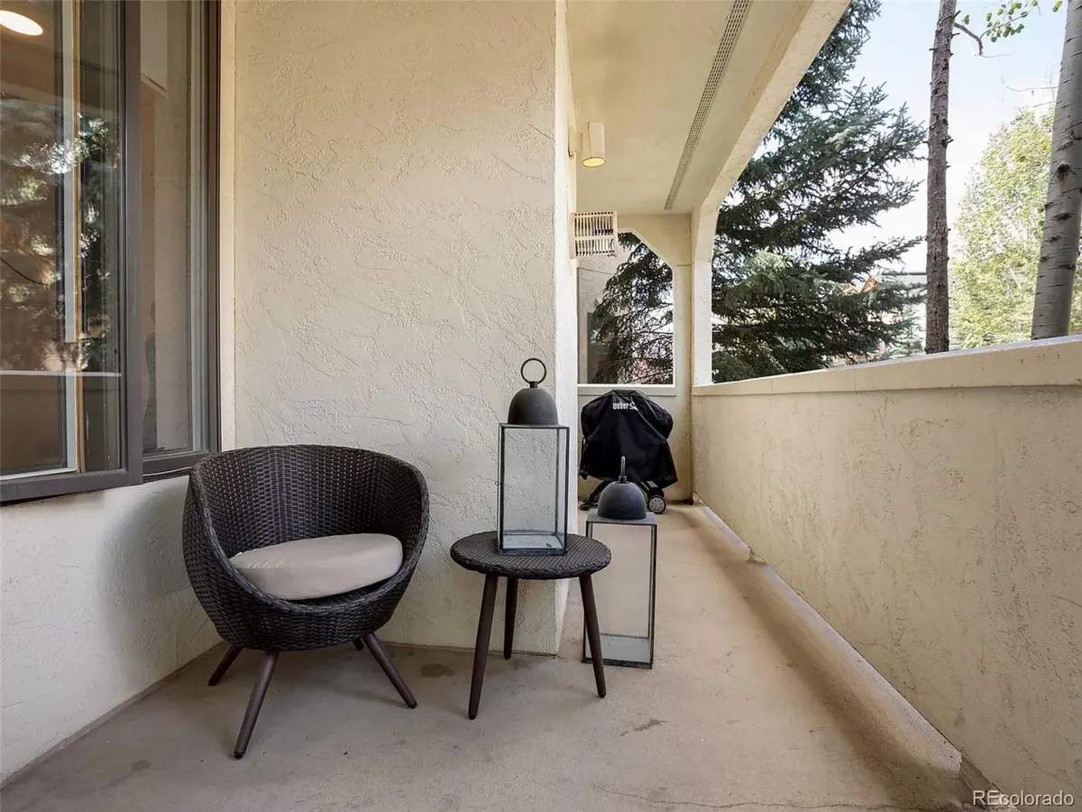A cozy covered balcony with a wicker chair, small table, and lanterns, set against a backdrop of lush trees. The space exudes tranquility and invites quiet moments of relaxation or socializing, with enough room to enjoy a cup of coffee or an evening drink while taking in the natural surroundings.