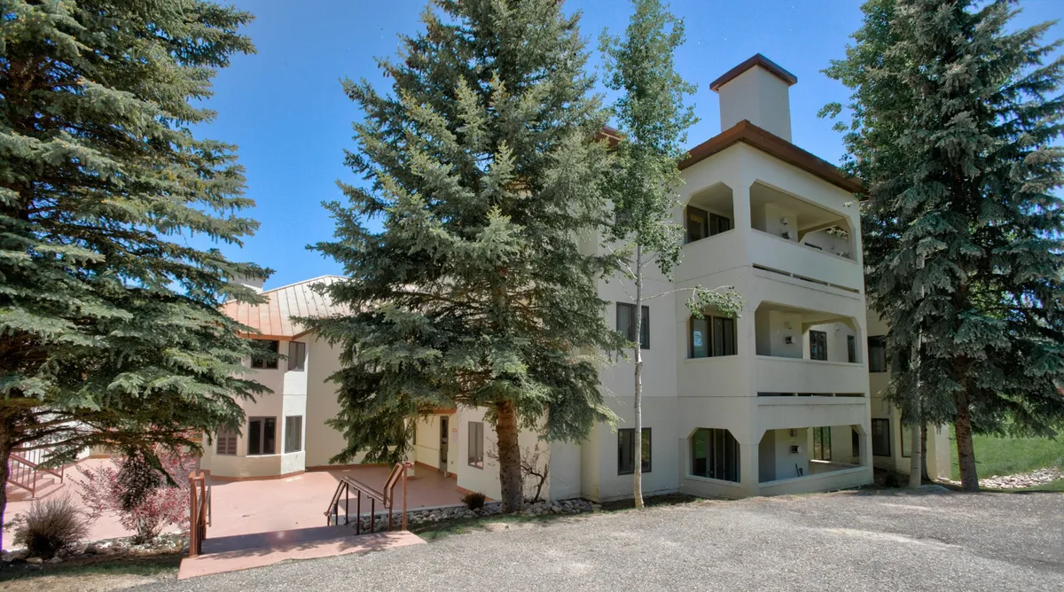 This exterior image captures the entrance to the multi-story building, surrounded by tall evergreen trees and a welcoming set of stairs. The greenery and architecture set the scene for the property's resort-style ambiance, hinting at both privacy and comfort within a natural setting.