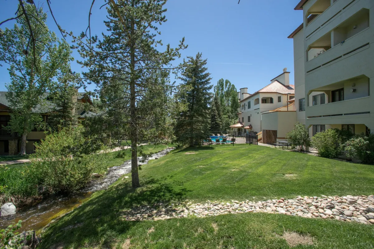 A lush green lawn slopes down to a meandering stream, bordered by trees and featuring a pool area with umbrellas. The scene is serene, showing spaces where guests can relax, read, or sunbathe, blending leisure with the natural surroundings.