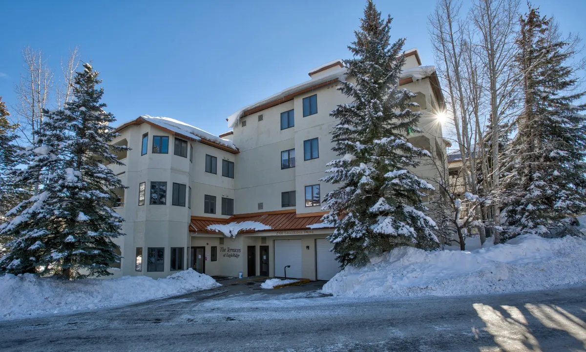 This exterior image shows the building facade, surrounded by snow-dusted trees under a crisp blue sky. The scene is vibrant and lively, hinting at winter fun and mountain adventure, with space that could easily be near ski lifts or shuttle pickup.