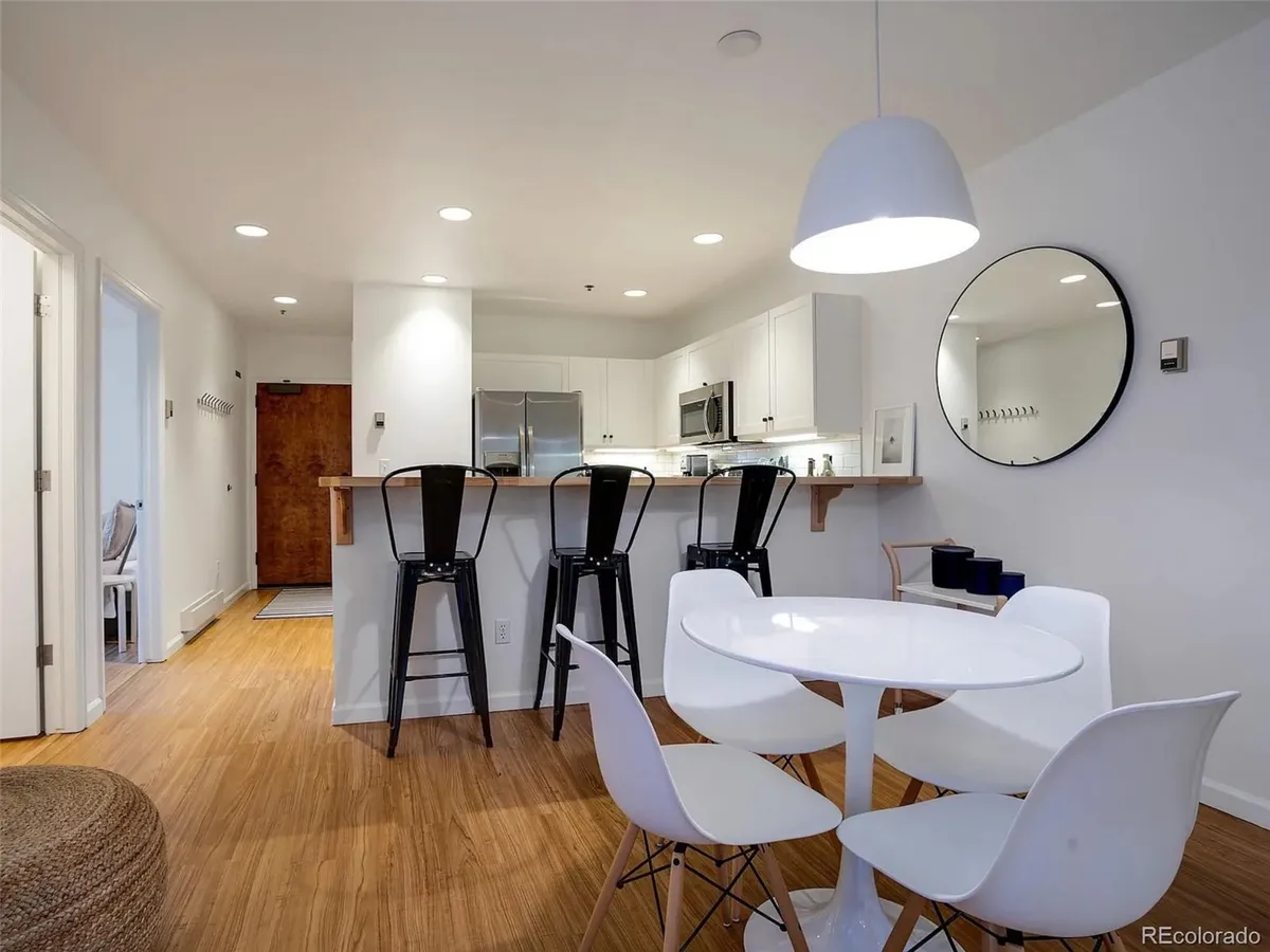 A modern, inviting kitchen and dining area with a large dining table, bar stools at a kitchen island, and a mirror reflecting the space. The overall mood is bright and social, ideal for shared meals or relaxing mornings.